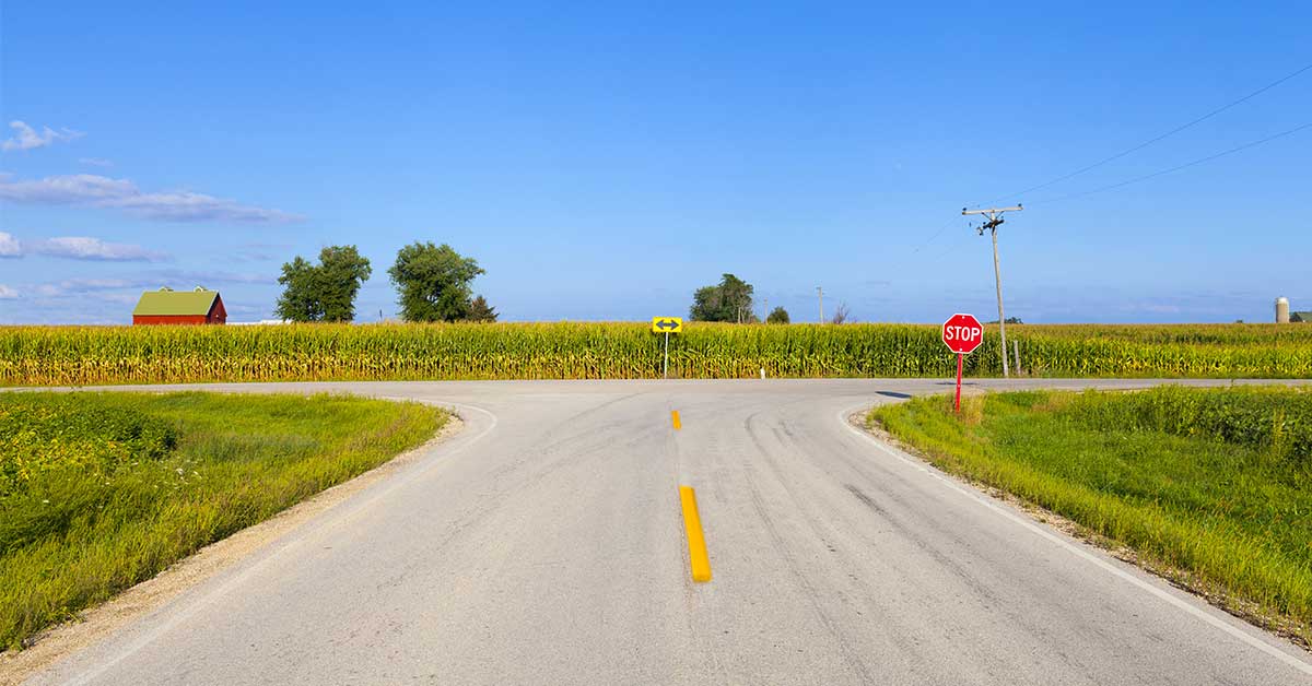 A rural intersection with a stop sign, yellow arrow sign, and cornfields on both sides under a clear blue sky invites intuitive reading of the landscape. A red barn and silo are visible in the distance.