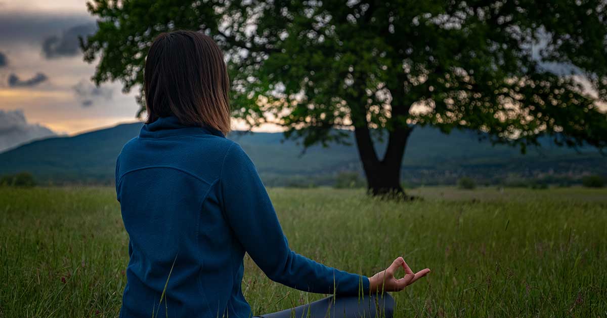 A person displaying empath traits sits cross-legged on grass, meditating outdoors at dusk and facing a tree, with mountains and clouds in the background.