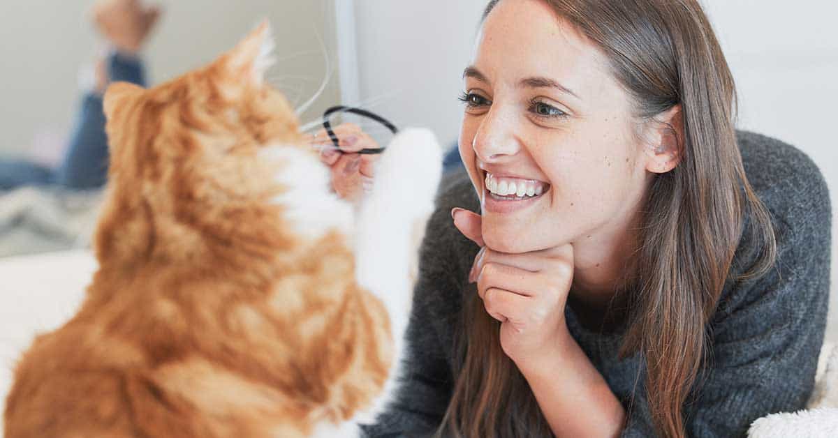 A woman smiles while facing an orange and white cat indoors, as if she can talk to animals.