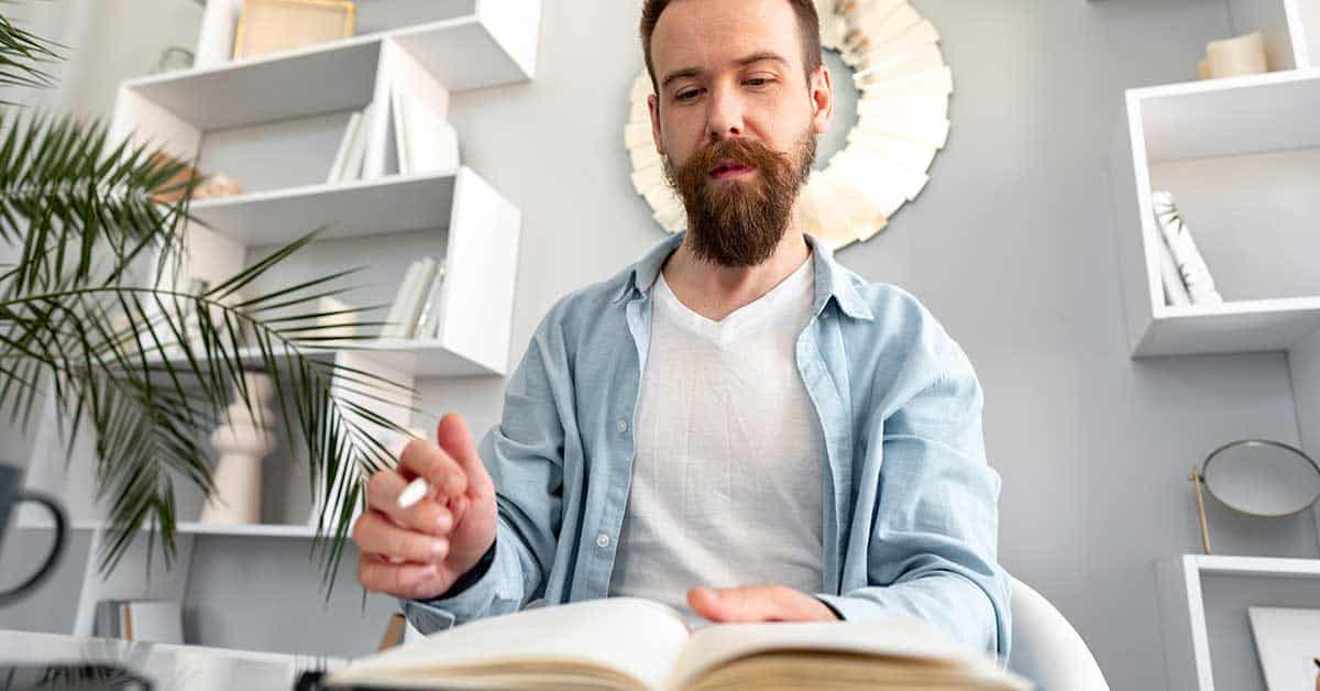 Bearded man in a light blue shirt sitting at a desk, reading an open book and holding a pen, as if preparing for intuitive readings, with shelves and a round mirror in the background.