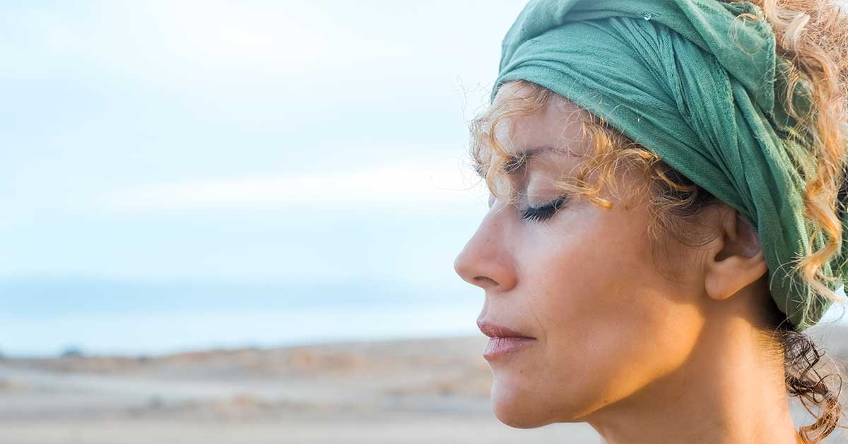 Woman with curly hair and a green headscarf stands outdoors with her eyes closed, facing left, as if embracing an intuitive reading in a serene, natural setting.