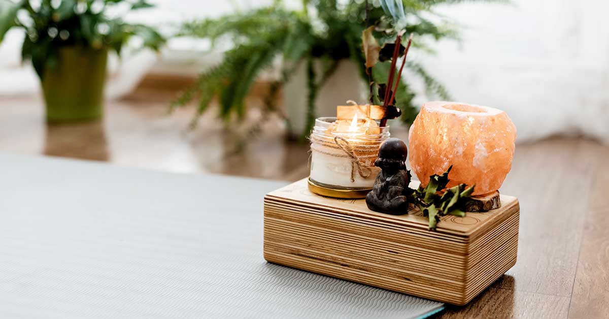 A yoga mat beside a wooden tray holding a candle, Himalayan salt lamp, small Buddha statue, and greenery sets the scene for an intuitive reading session, with calming potted plants in the background.