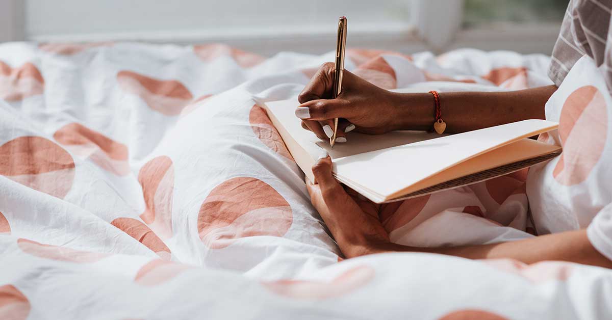 A person sits on a bed with patterned bedding, trusting their intuition as they write thoughtfully in an open notebook with a pen.