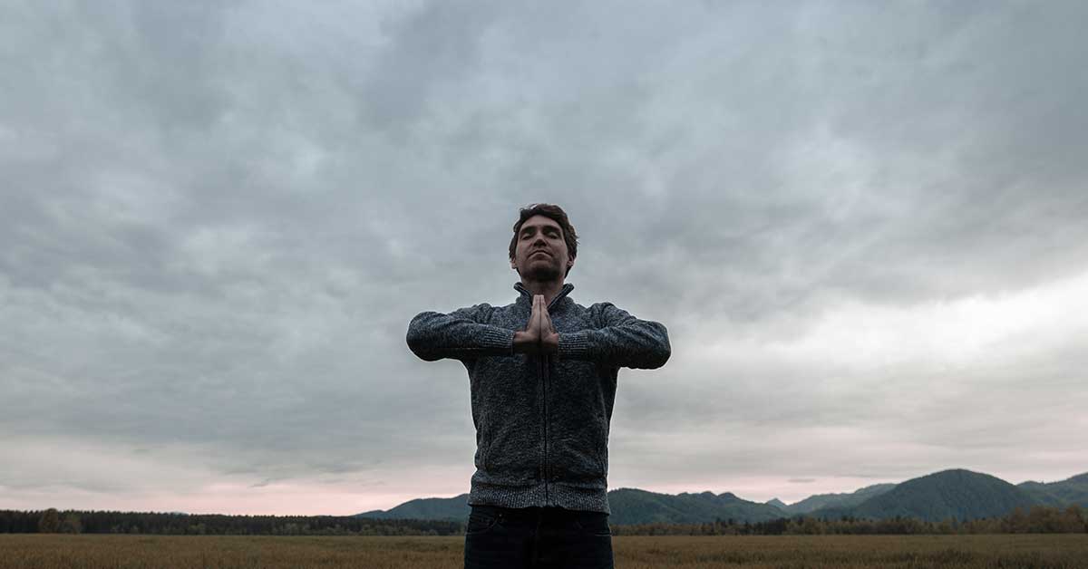 A person stands outdoors with hands pressed together in a prayer position, radiating an intuitive calm amid mountains and a cloudy sky in the background.