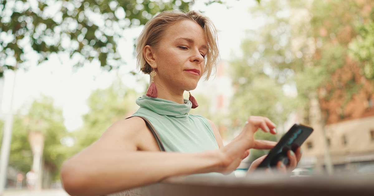 A woman sits outdoors on a bench, engaged in intuitive reading on her smartphone, with trees and buildings blurred in the background.