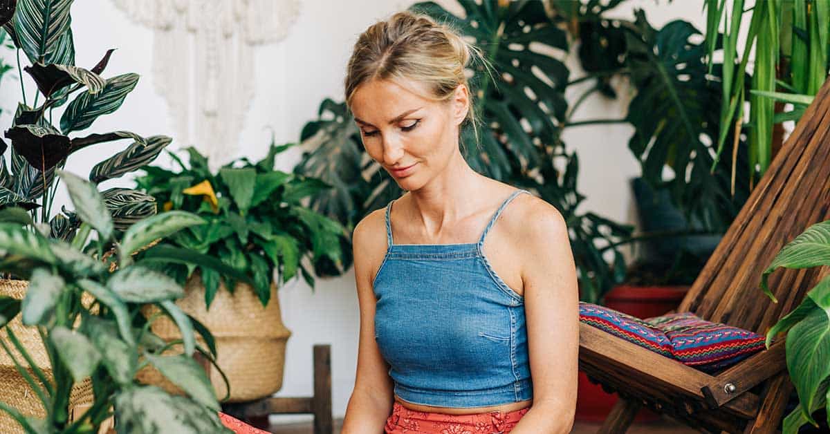 A woman in a blue sleeveless top sits among various potted plants, looking down with a calm expression, as if finding peace of mind amidst an Intuitive readings