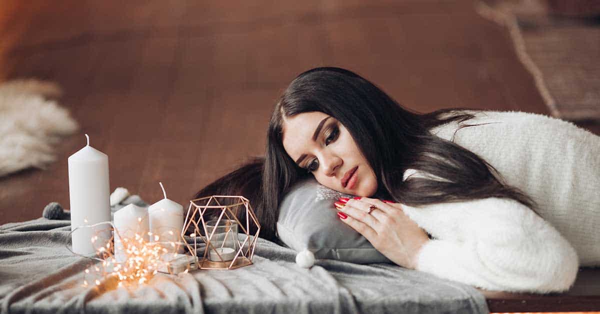 Woman with long dark hair lies on a gray cushion, surrounded by candles, string lights, and decorative items on a cozy blanket—creating a serene space for reflection, grief, and intuitive reading.