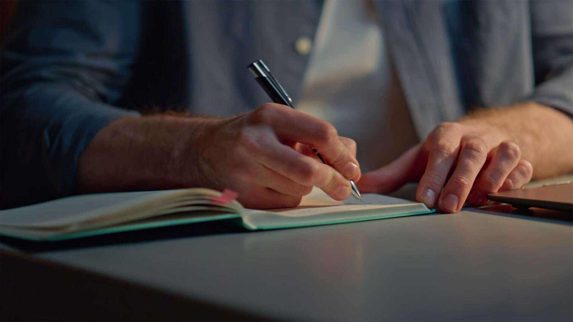 A person writes in an open notebook with a pen while sitting at a desk, surrounded by reading materials.