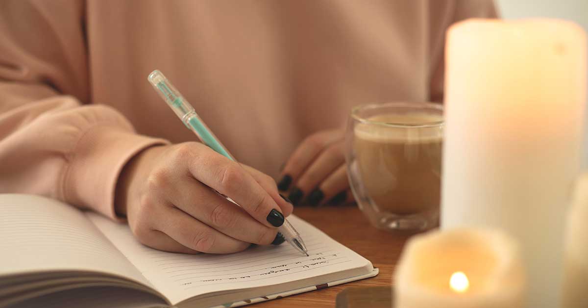 Person writing in a notebook with a pen, holding a cup of coffee, and lit candles on the table—setting the mood for intuitive guidance and inspired journaling.