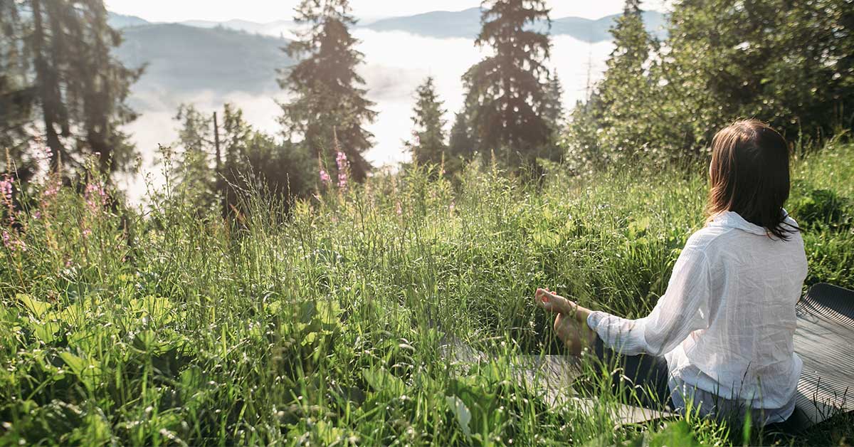 A person sits cross-legged on a mat in a grassy area, surrounded by trees and wildflowers, facing a misty landscape, embracing an intuitive connection with nature.