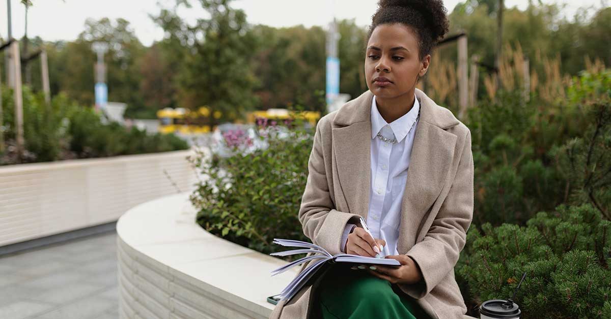 A woman sits outdoors on a curved white bench, immersed in intuitive reading as she writes in a notebook. She wears a beige coat and white shirt, with greenery and blurred yellow buses in the background.