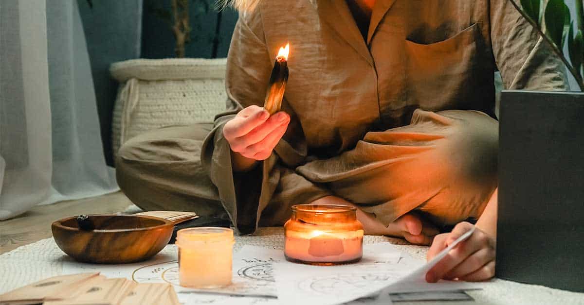 A person in brown pajamas sits cross-legged on the floor, holding a lit smudge stick near candles and papers with drawings, possibly engaging in a ritual, meditation, or intuitive readings.