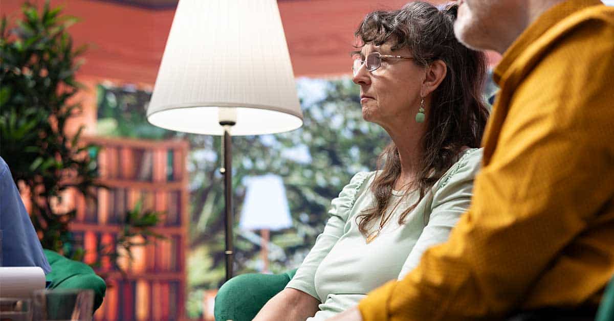 Two adults sit side by side, engaged in conversation with a third person off-frame, as if receiving intuitive readings. A lamp and bookshelf are visible in the background.