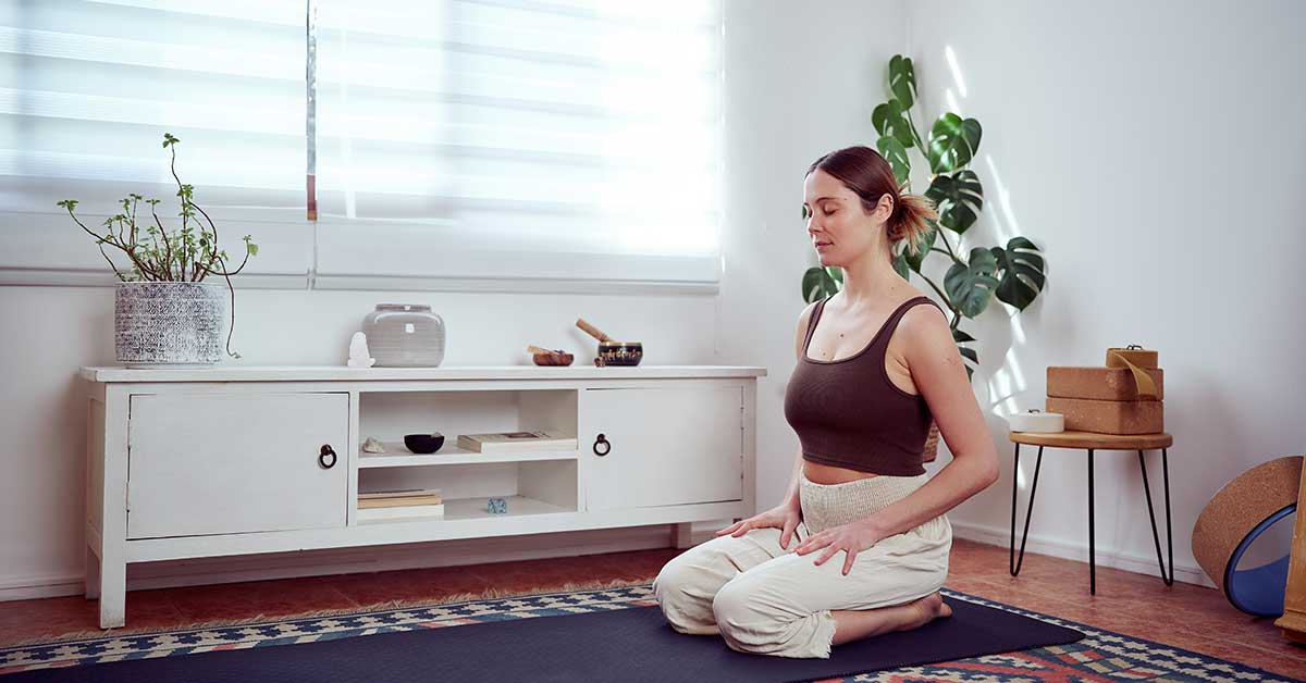 A woman sits on her knees in a meditative pose on a yoga mat in a bright, minimalistic room with plants and yoga props, preparing for an intuitive reading.