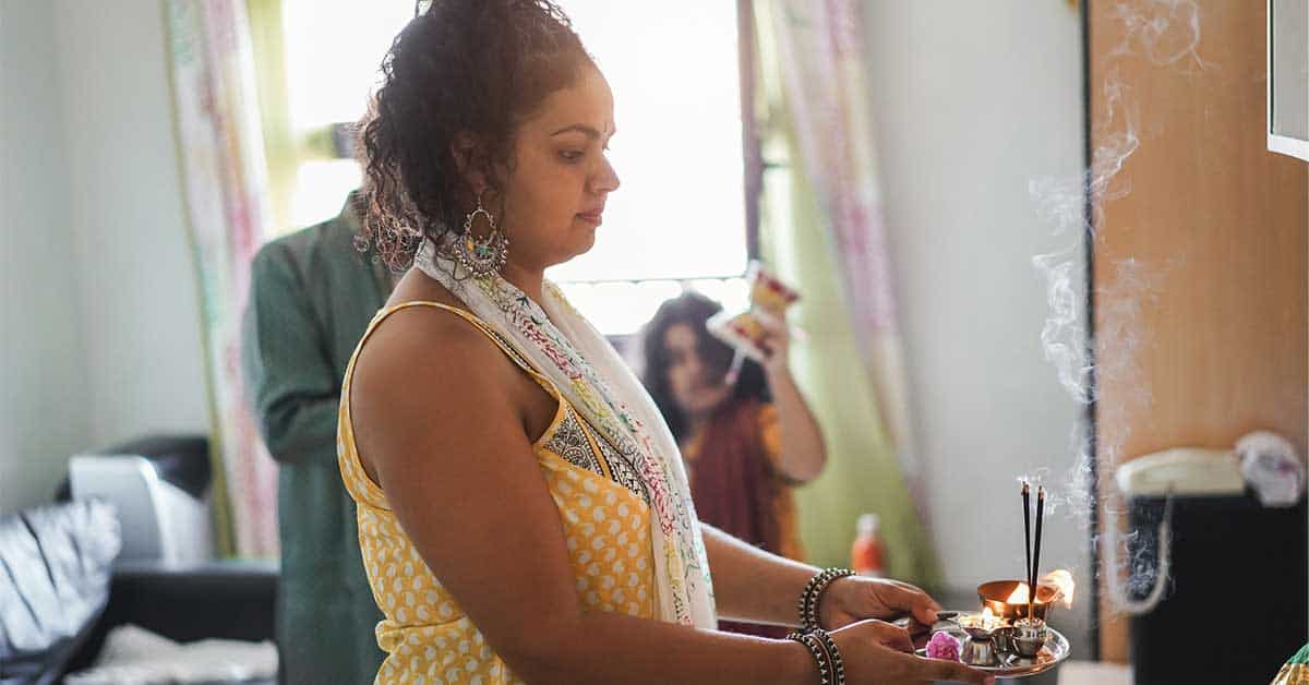An intuitive woman in traditional attire performs a puja ritual indoors, holding a plate with incense and offerings as smoke rises, while others observe in the background.