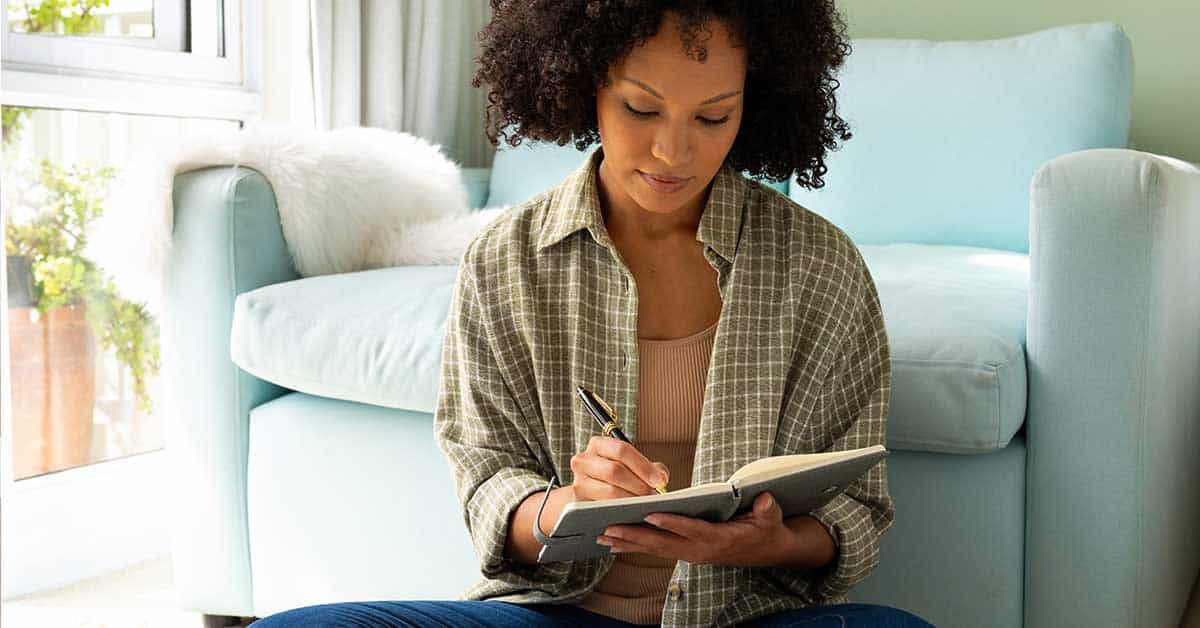 A woman sits on the floor in front of a sofa, writing in a notebook with a pen, as she reflects on her inner wisdom to guide your journey.