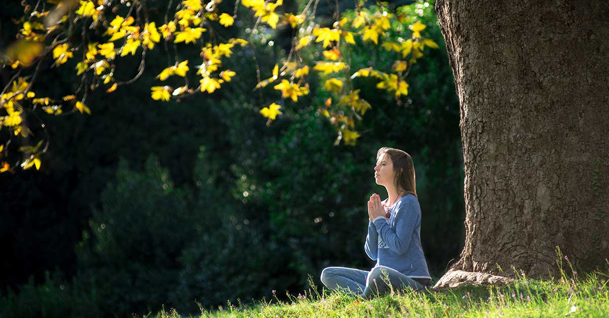 A woman sits cross-legged on grass under a large tree, hands clasped together and eyes closed, as she taps into her intuition amid sunlight and greenery.