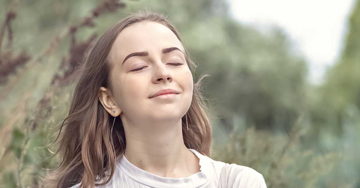 A young woman with long hair stands outdoors with her eyes closed and a slight smile, surrounded by greenery, as if finding peace after intuitive readings.