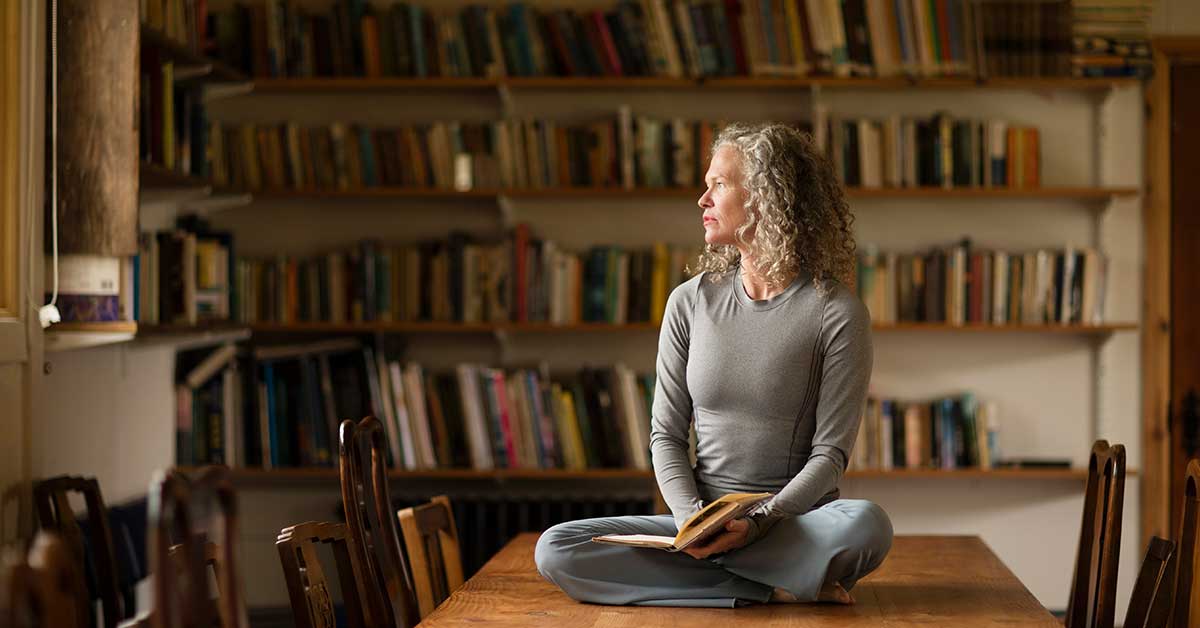 A woman sits cross-legged on a wooden table, holding an open book for an intuitive reading, as she looks out a window in a library lined with bookshelves.