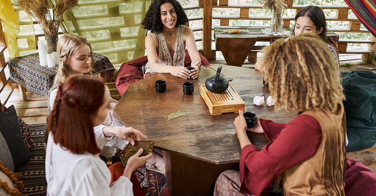 Five people sit around a wooden table with cups and a teapot, engaging in conversation and an intuitive reading in a cozy, bohemian indoor setting.