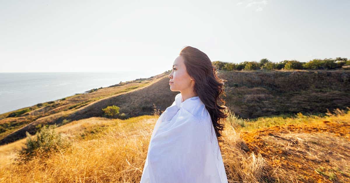 A woman draped in a white cloth stands outdoors on a grassy hillside, facing the ocean under a clear sky, as if guided by intuition.