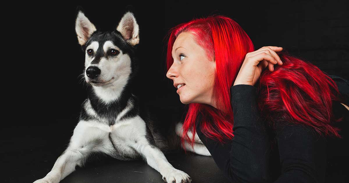 A woman with bright red hair lies beside a loyal black and white dog on a dark background, embodying her role as an animal communicator who understands the unique bond and difference between pet and human.