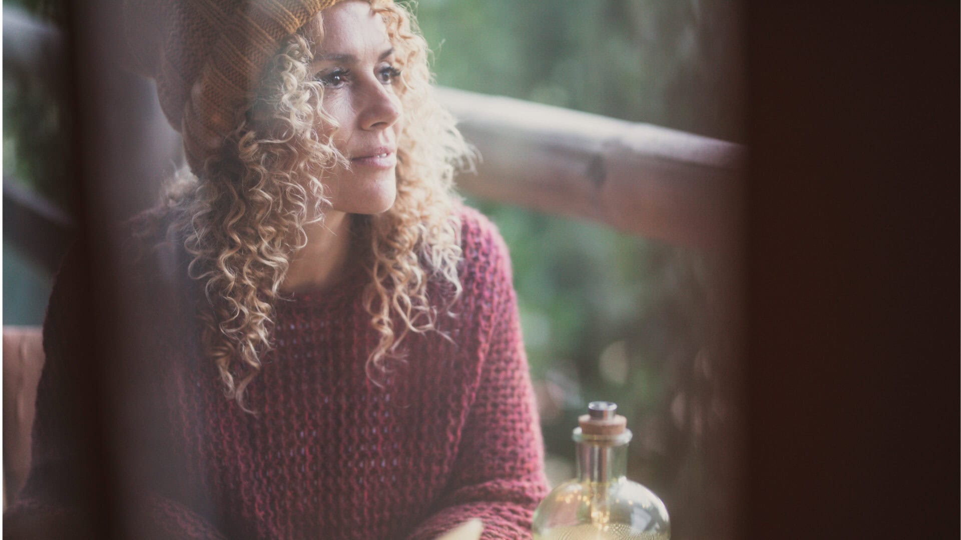 An intuitive person with curly hair in a knit hat and sweater sits by a window, gazing outside thoughtfully, with a glass bottle nearby.