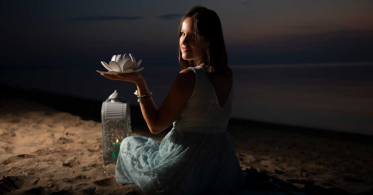 A woman in a white dress sits on a sandy beach at dusk, holding a lit lotus-shaped lamp, seeking hidden opportunities.