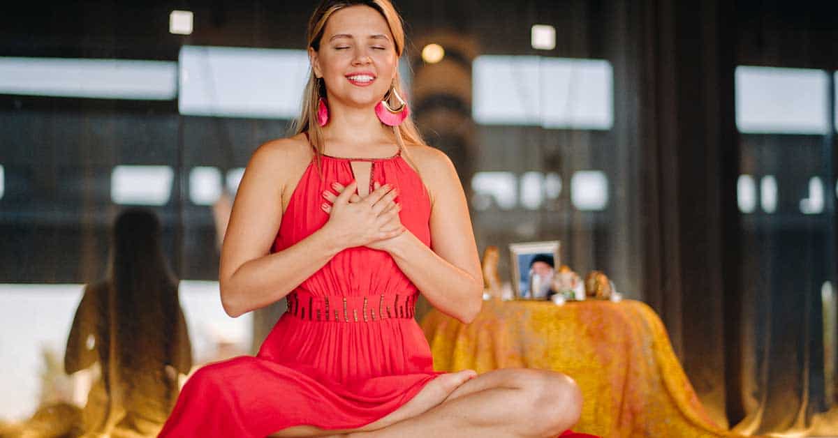 A woman in a red dress sits cross-legged indoors, hands on her chest, eyes closed, embodying a calm expression as she seeks spiritual guidance at her career crossroads. A small table with objects rests serenely in the background.