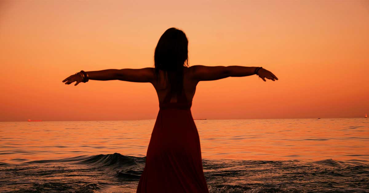 Silhouette of a person with outstretched arms facing the ocean during sunset, embracing the serenity of ancestral wisdom, akin to modern intuitive life coaching that inspires personal growth and transformation.