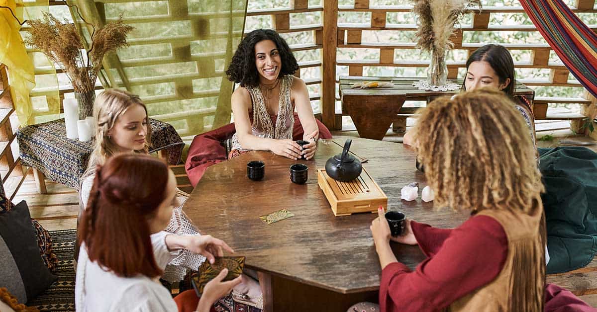 A group of five people sit around a round wooden table, sharing tea and engaging in intuitive readings in a cozy, decorated room.