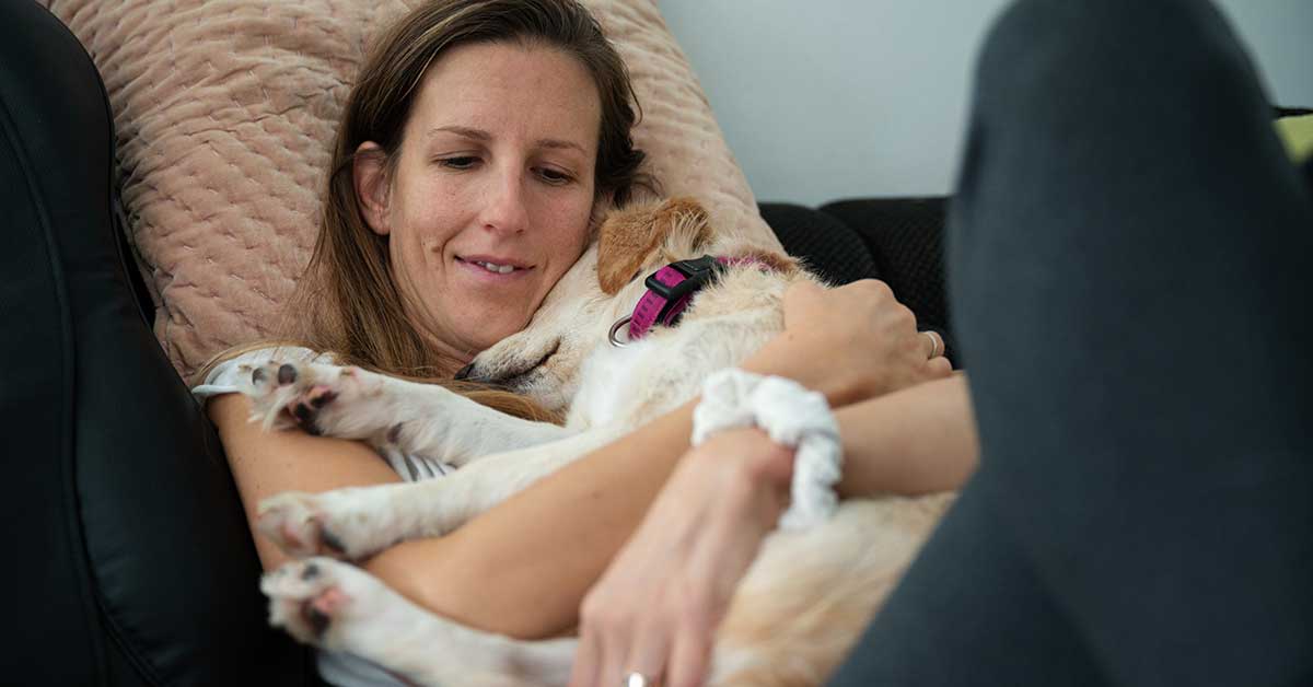 A woman is lying on a couch, smiling, and holding her small dog closely as they both rest, embodying the joy and tranquility many pet owners cherish.