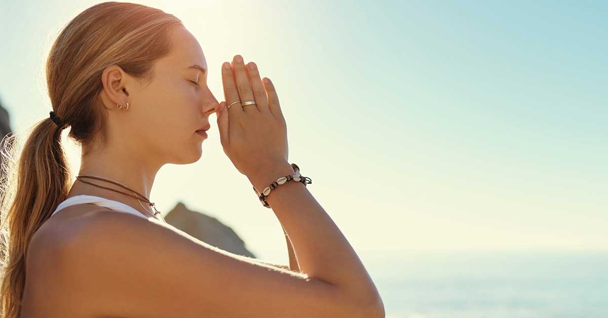 A woman with closed eyes holds her hands in prayer pose, standing against a bright sky, as if to protect her energy.