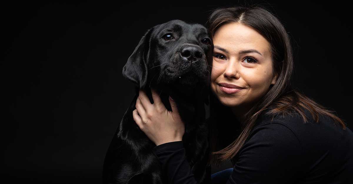 A woman with dark hair in dark clothing, embodying the warmth of pet support, embraces a black Labrador Retriever against a black background.