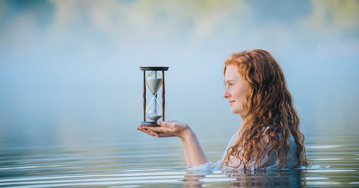 A woman with long red hair holds an hourglass, embodying divine timing as she stands waist-deep in calm water, surrounded by mist.