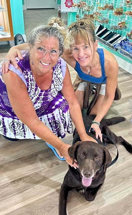 Two women kneel on a wood floor, smiling at the camera, with their hands on a large chocolate labrador dog lying between them in an indoor setting.