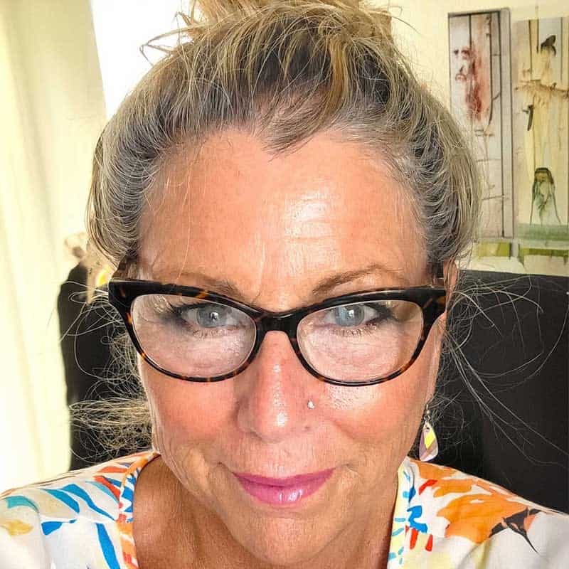 A woman with glasses and a nose piercing, wearing a colorful floral top, looks at the camera with a slight smile while indoors.